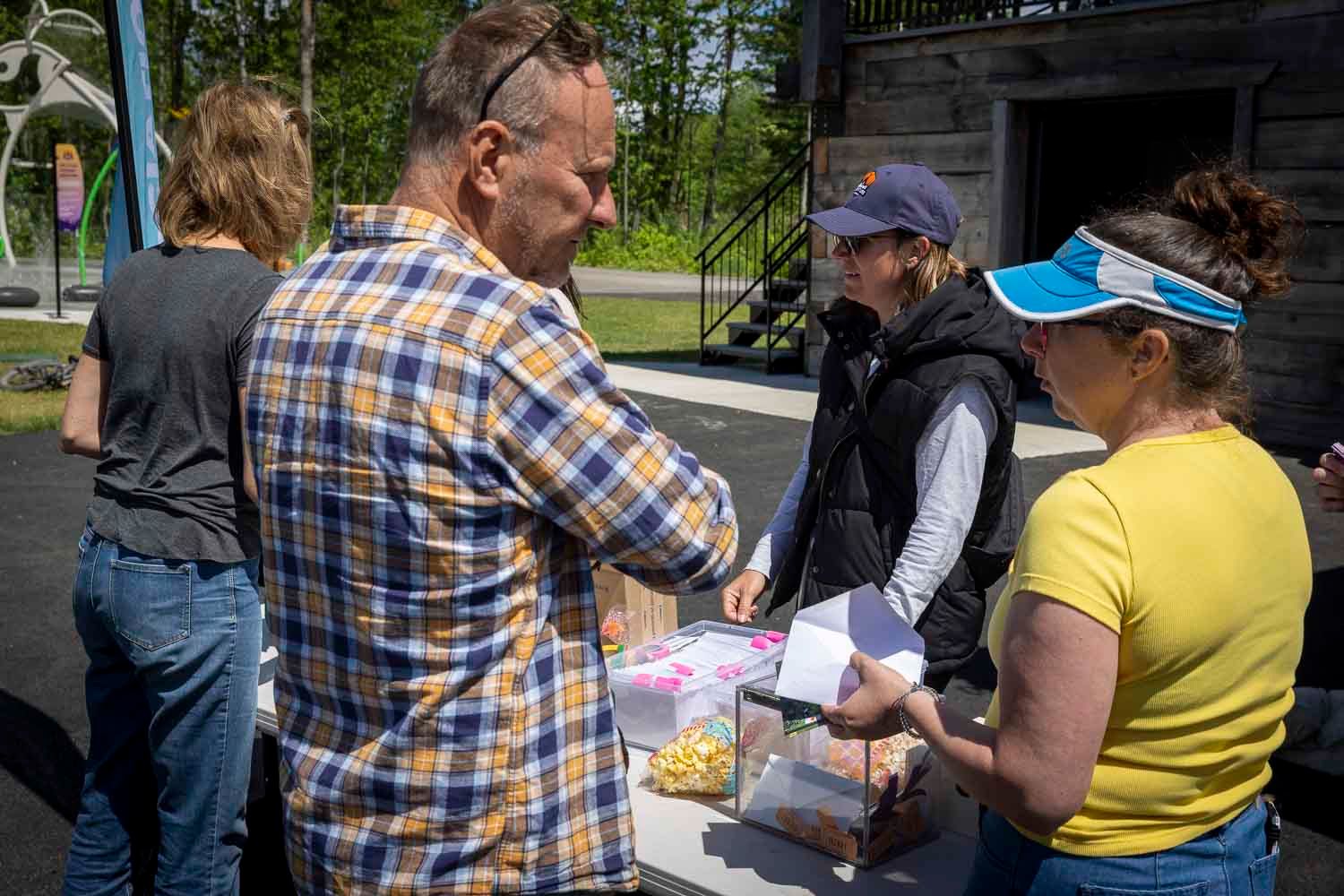 People gathered outdoors exchanging items at a sunny community event near a wooden building.