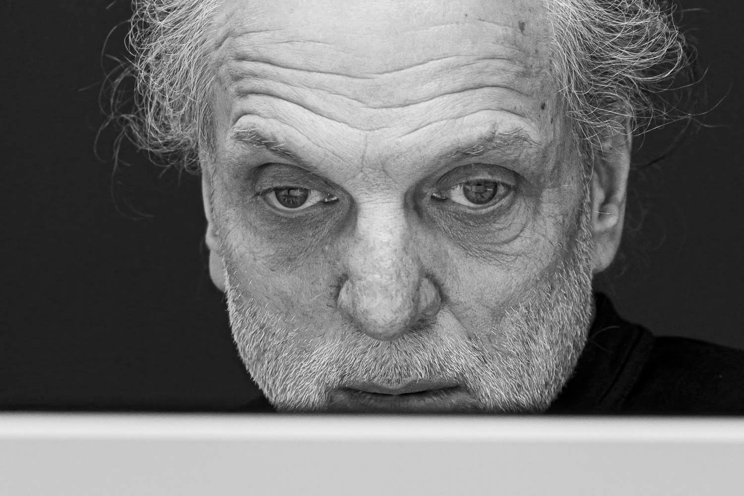 Black and white portrait of an elderly man with a beard and intense gaze peering over a white surface.