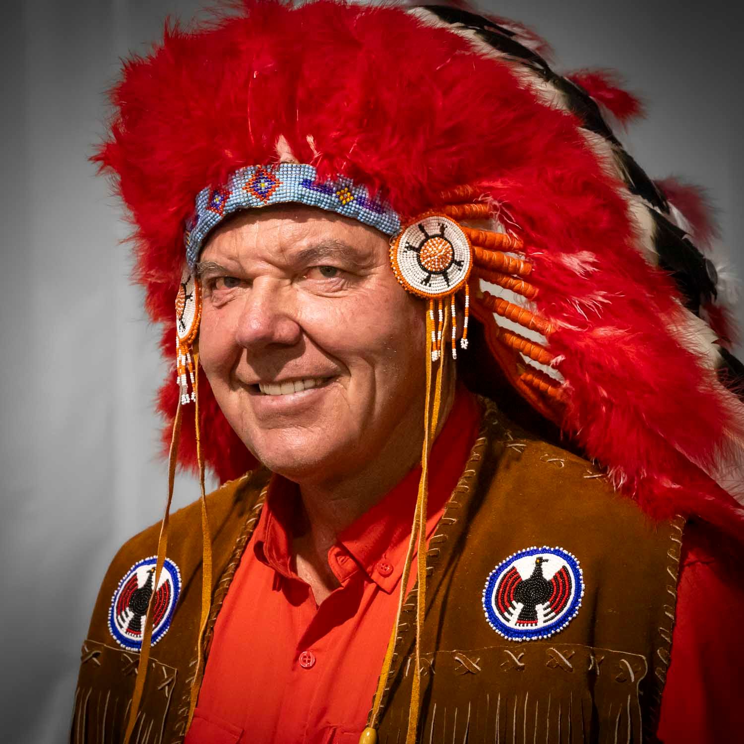 Man wearing traditional Native American headdress with red feathers and beaded details, dressed in fringed leather vest.