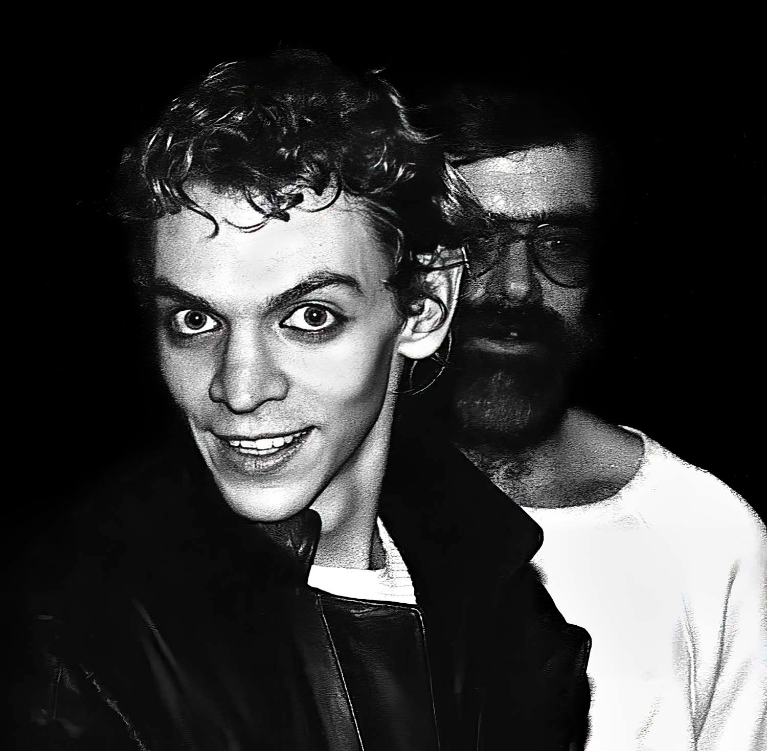 Black and white photo of two men, one young with wide eyes smiling, another bearded man with glasses behind him.