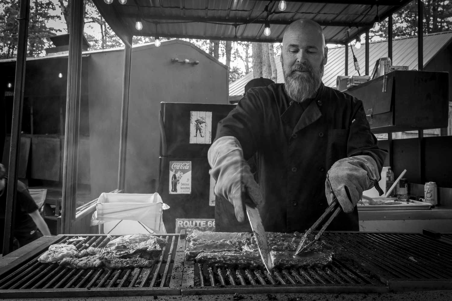 Black and white photo of a bearded man grilling meat outdoors at a BBQ stand using tongs.