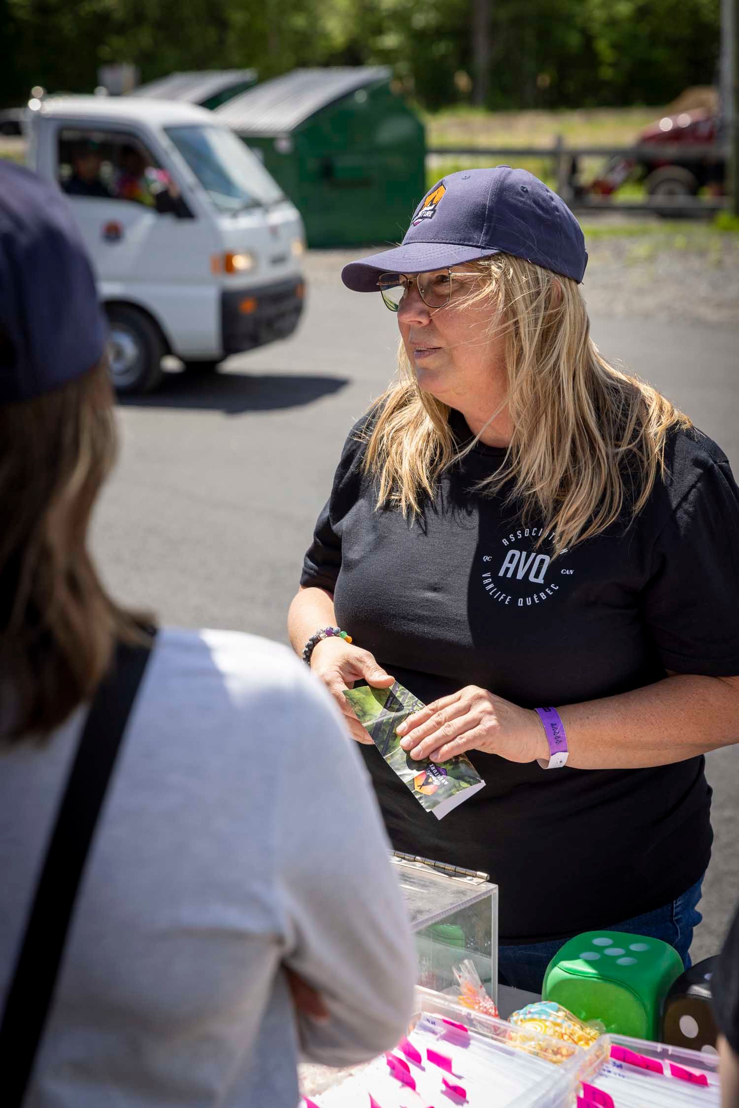Woman in black AVO shirt and blue cap handing items at an outdoor vendor table on a sunny day.