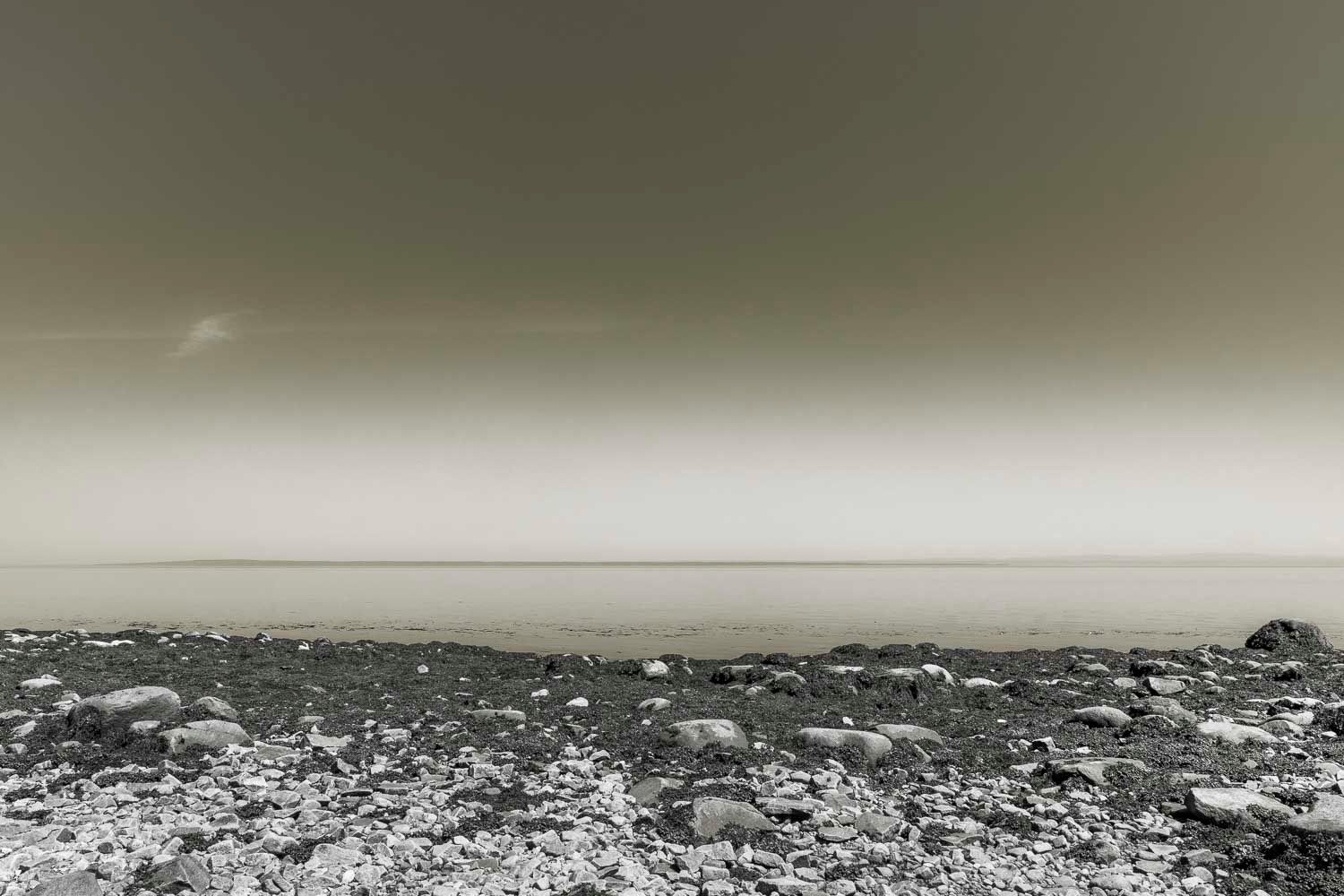 A desolate rocky shoreline meets a calm misty sea under an overcast sky in black and white.