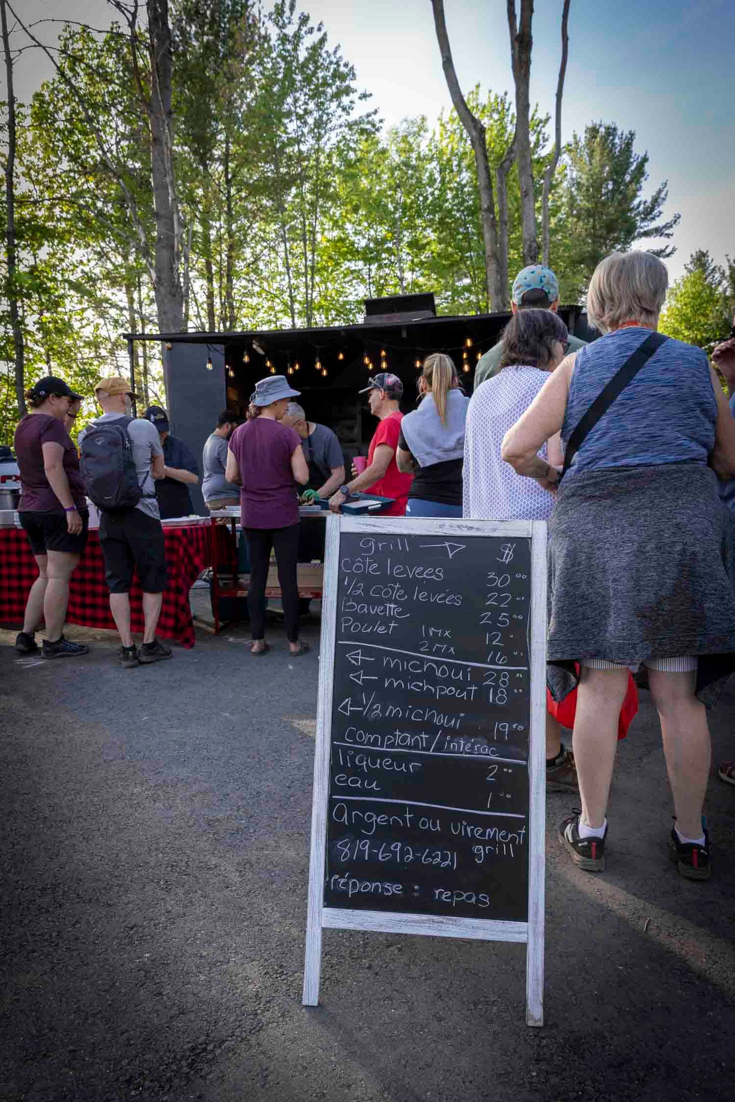 Outdoor market queue with chalkboard menu sign, people browsing food stalls surrounded by trees.