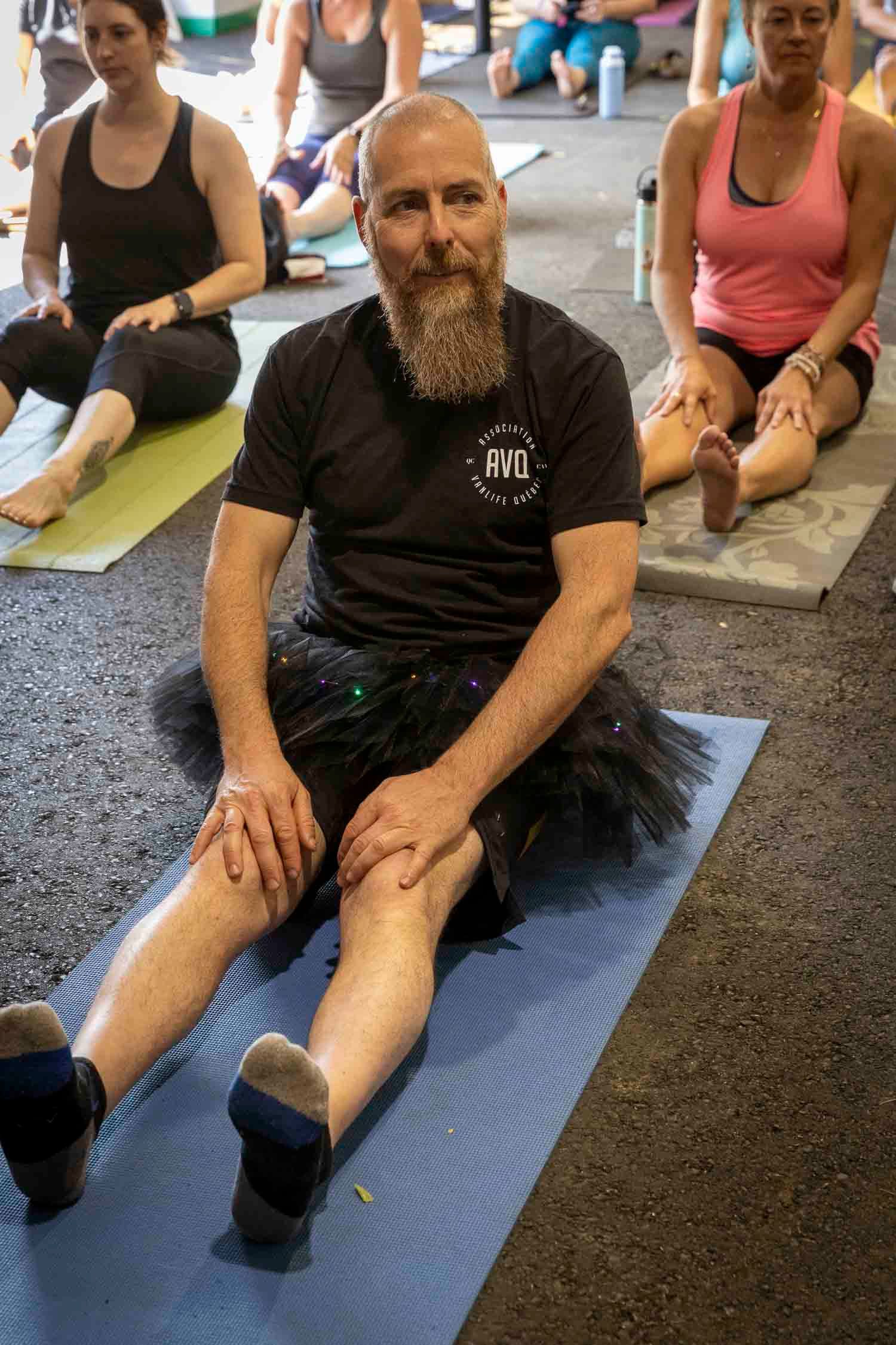 Bald bearded man sits on yoga mat wearing black tutu during group yoga class indoors.