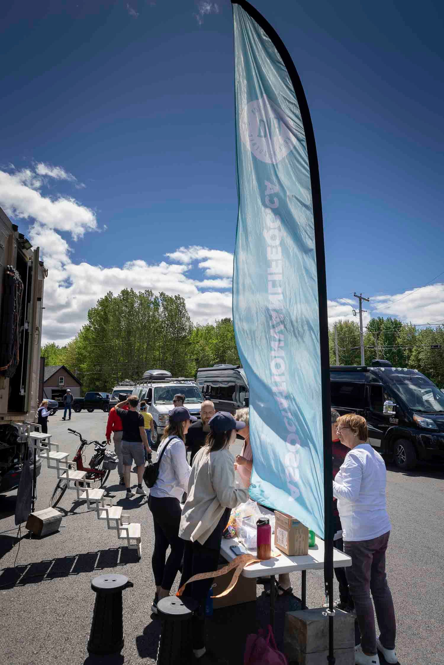 Outdoor event booth with a tall teal feather flag banner, people gathered around a table under a sunny blue sky.