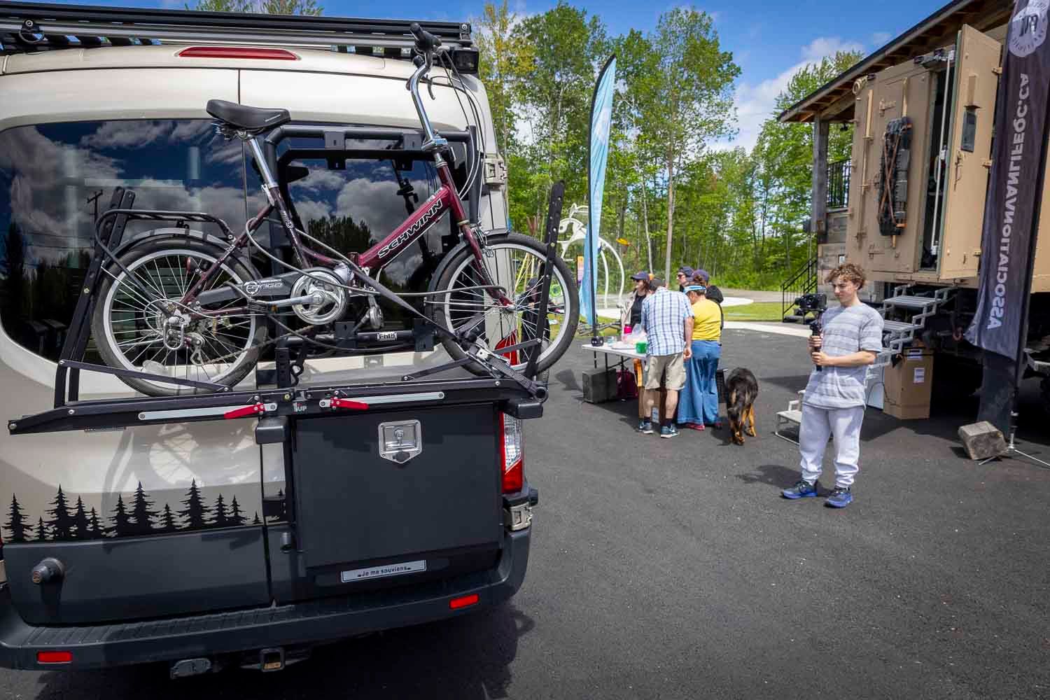RV with bike rack carrying a mountain bike at an outdoor camping expo with visitors and a dog nearby.