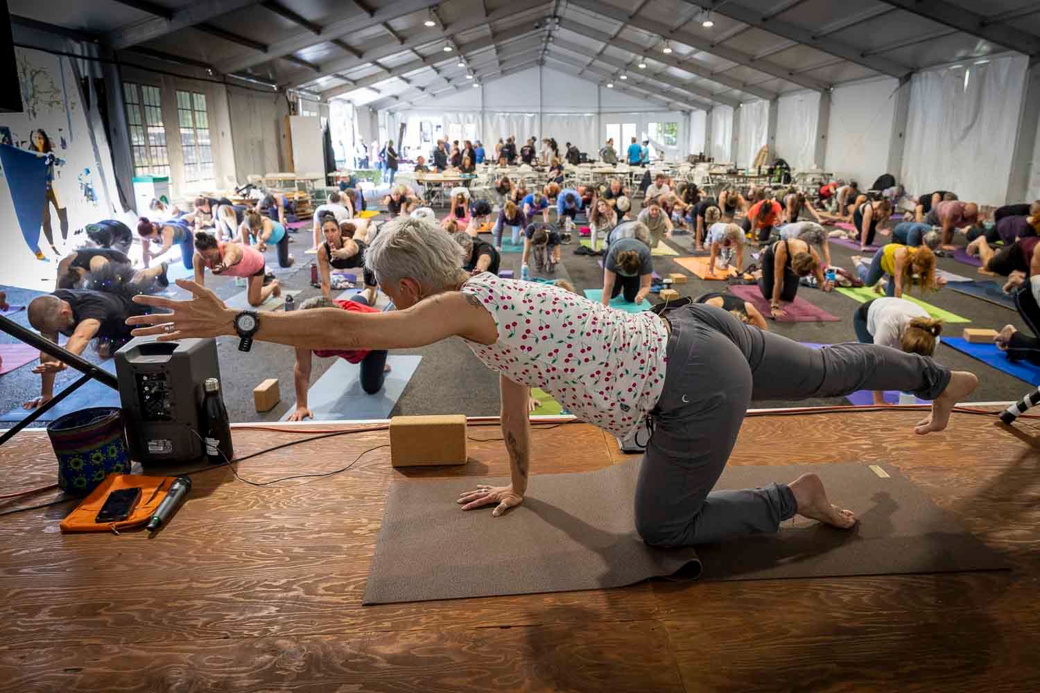 Yoga instructor demonstrating a tabletop pose on a mat before a large crowd in an indoor event space.