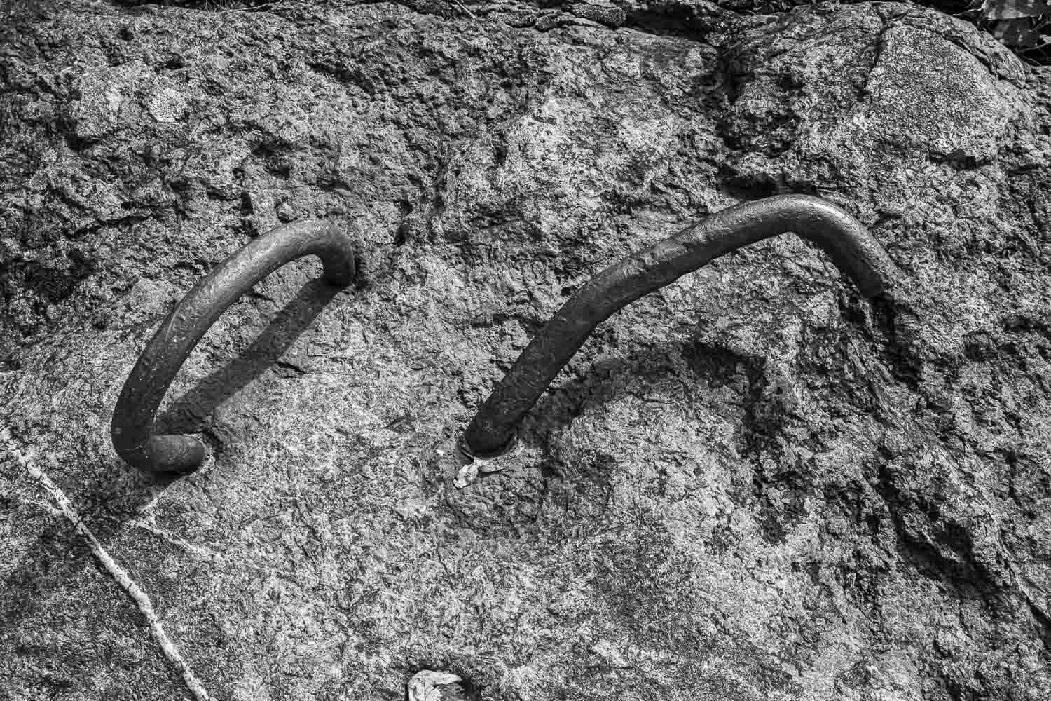 Black and white close-up of two rusty iron rings embedded into a weathered rock surface near a waterfront.