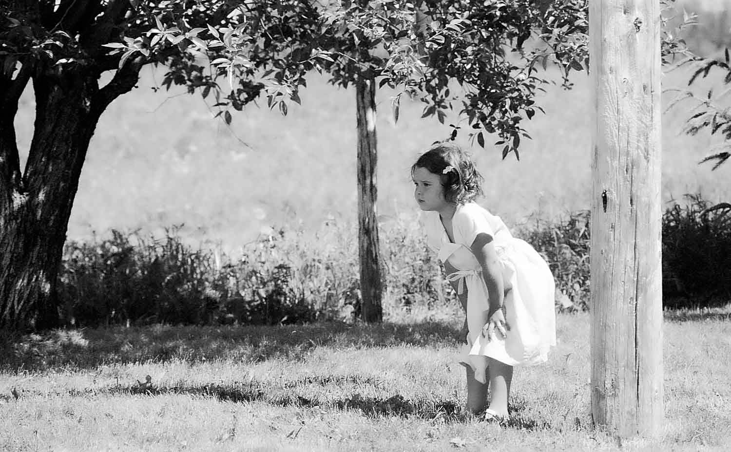 Jeune fillette dans un champs
A young girl in a white dress peeks around a wooden pole in a grassy yard with trees in the background.