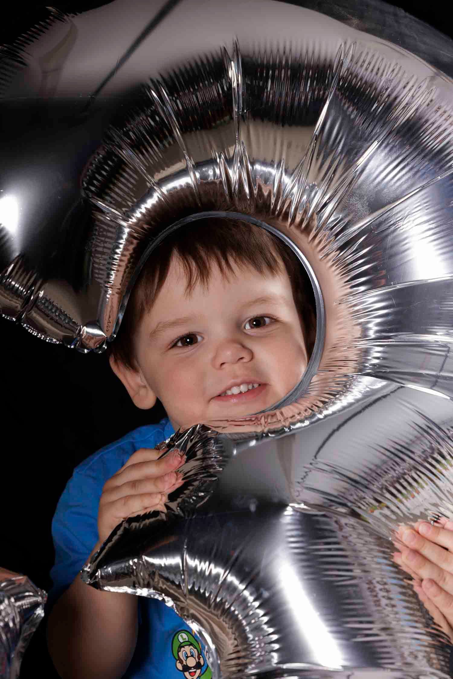 Happy toddler boy peeking through shiny silver number balloons at a birthday celebration.