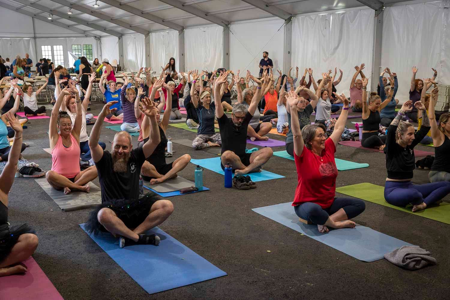 Large group yoga class with participants sitting cross-legged on colorful mats raising arms overhead in a tent venue.