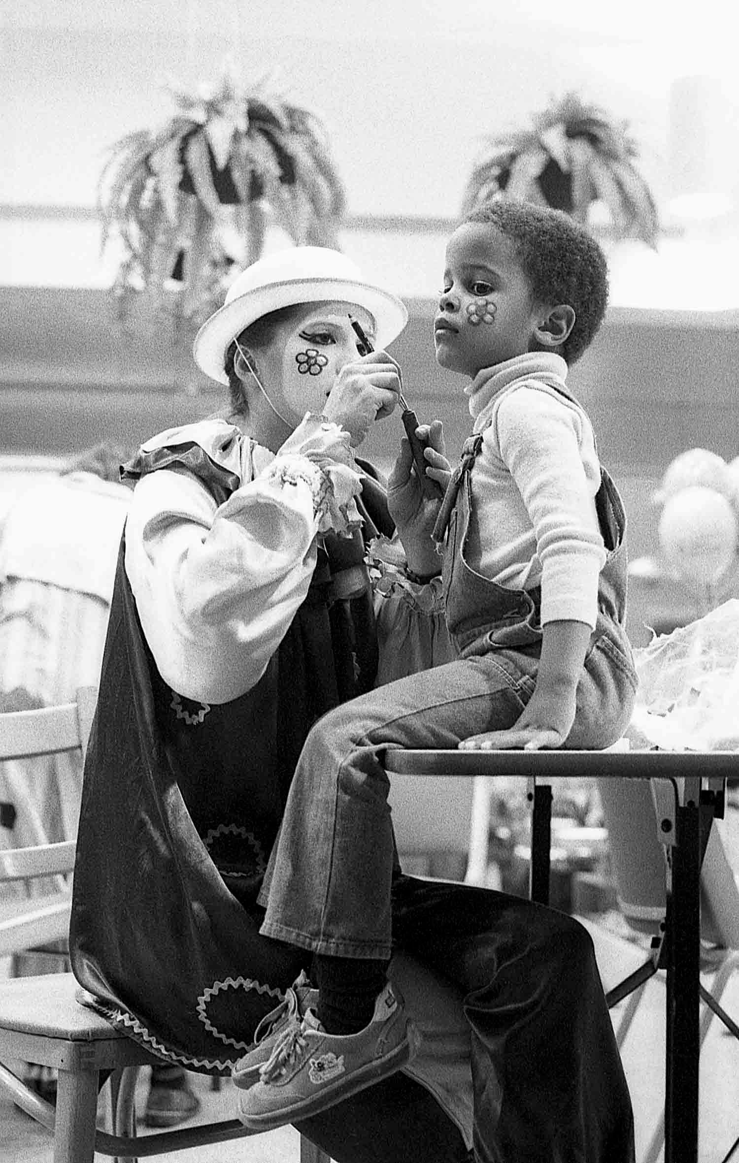 Black and white photo of a clown in costume painting the face of a young child seated on a table.
