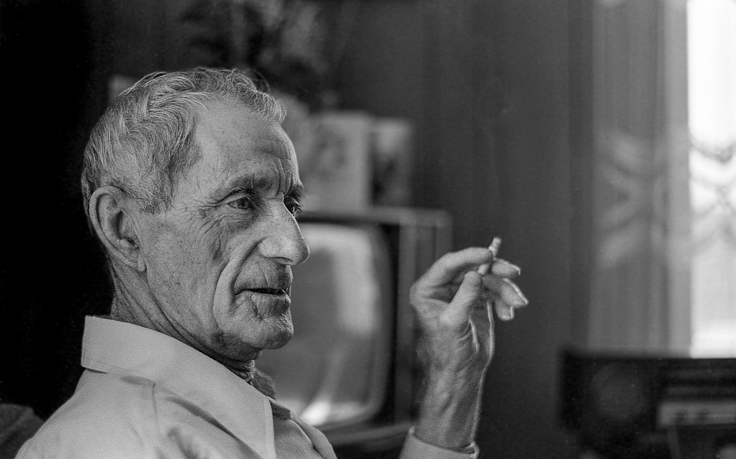 Elderly man in profile holding a cigarette, black and white portrait photo with vintage TV in background.