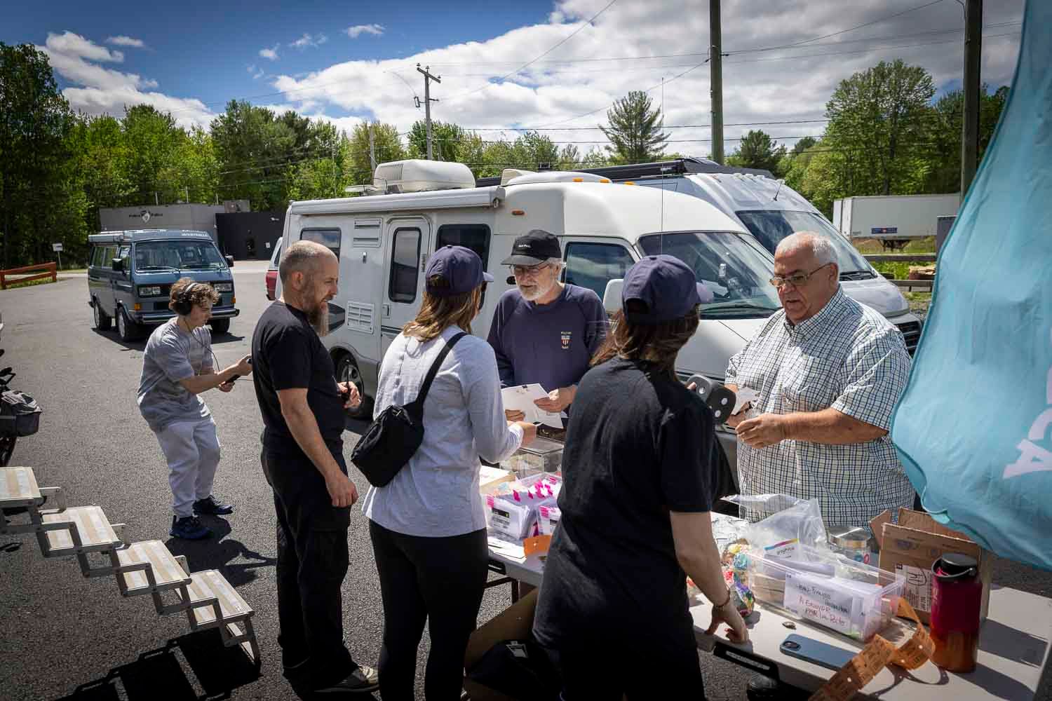 Group of people gathered around an outdoor food table near parked RVs and vans on a sunny day.