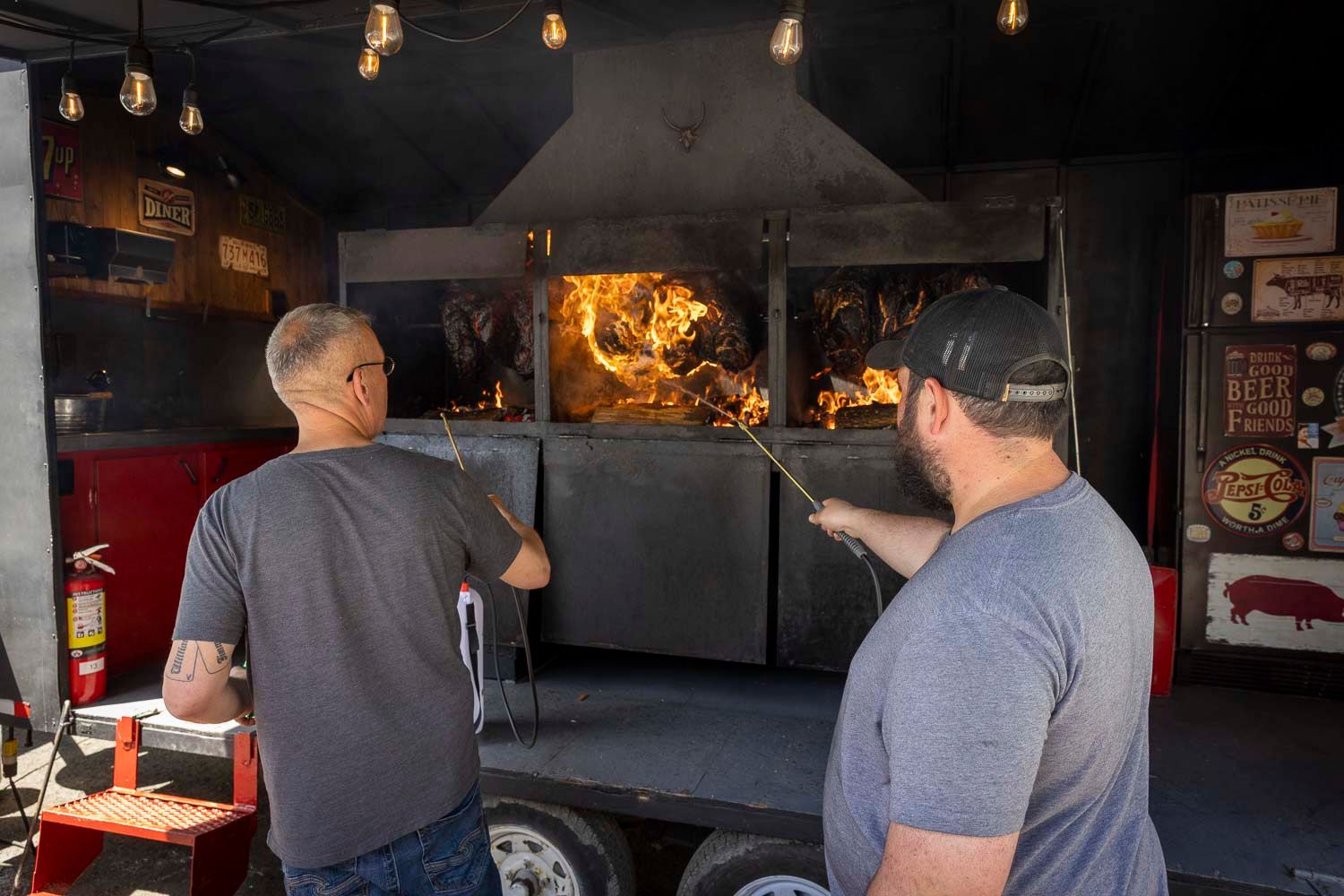 Two men tend to a wood-fired BBQ smoker with open flames inside a food trailer at an outdoor event.