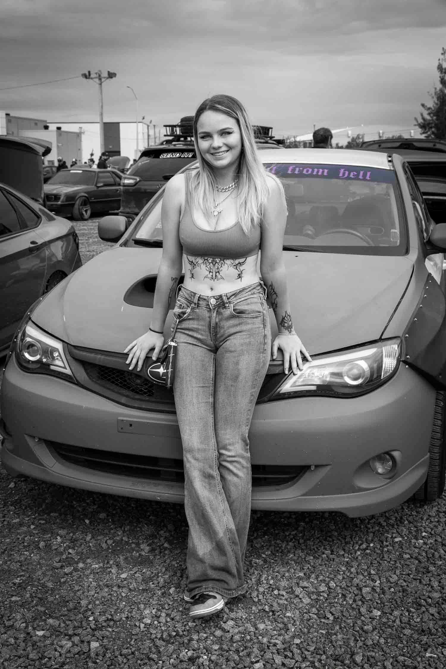Jeune femme et son auto, concours de beauté 
Woman in jeans posing in front of a Subaru car at an outdoor car meet in black and white photo.