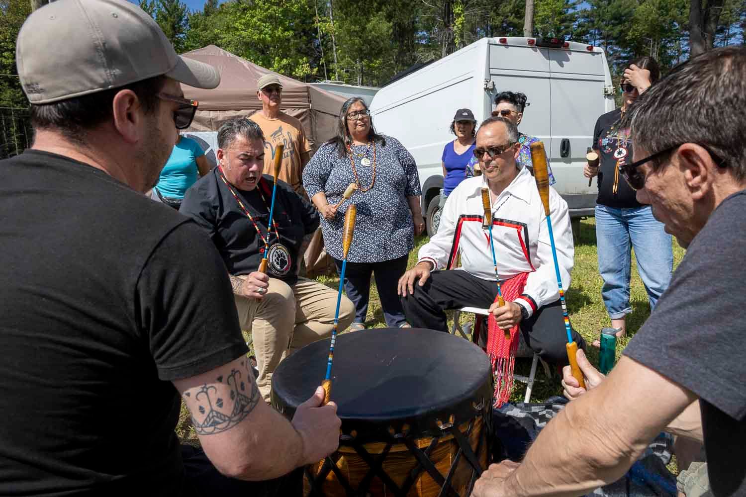 Group of people gathered outdoors around a drum during a traditional Indigenous drumming ceremony.