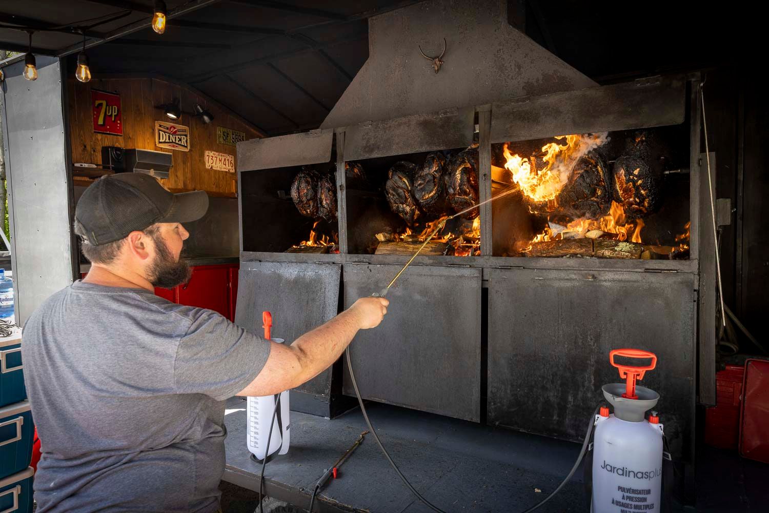 Man using a spray bottle to tend to meat cooking in a large outdoor BBQ smoker.