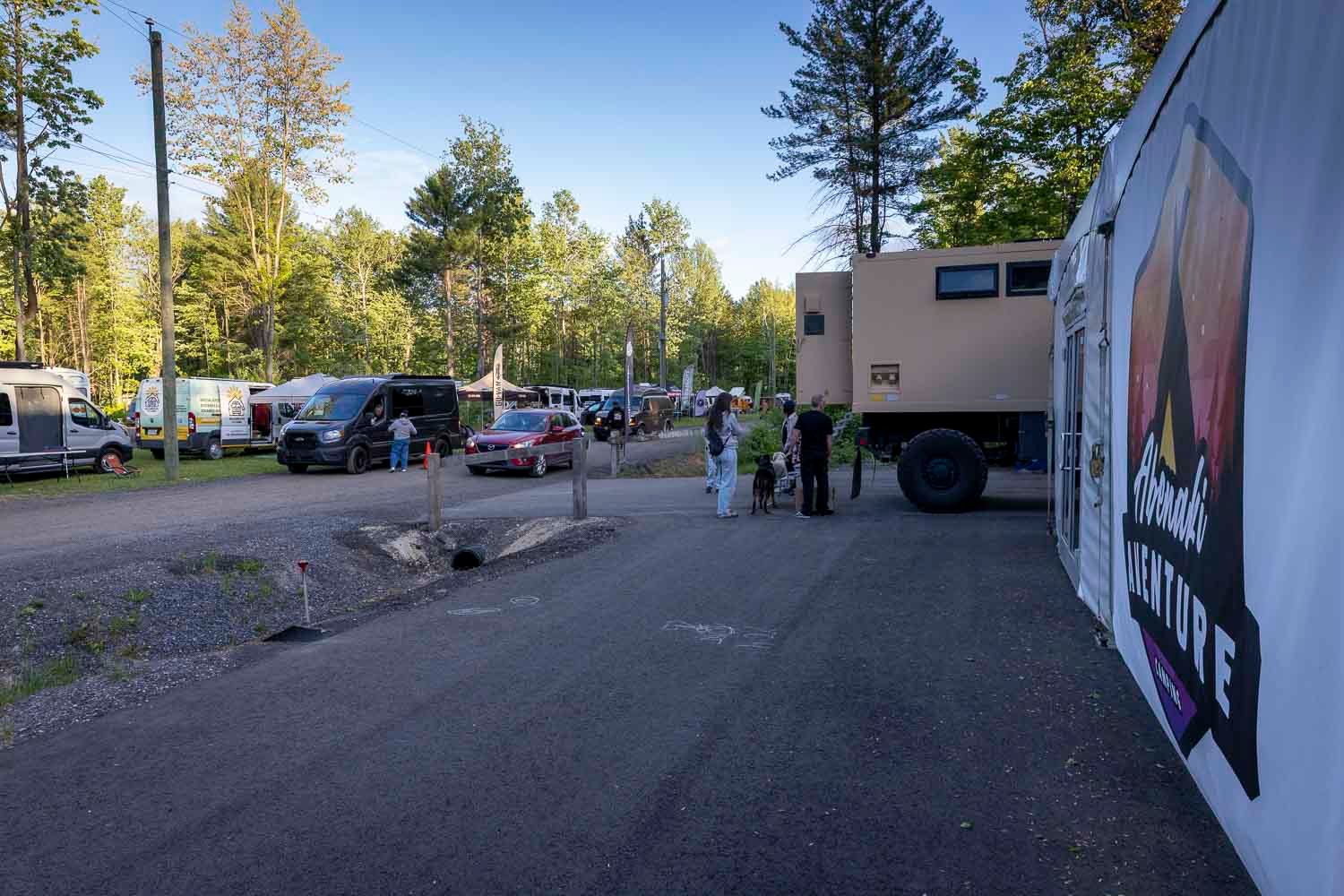 Outdoor campsite with vehicles, RV, and people gathered among tall trees on a gravel road at dusk.