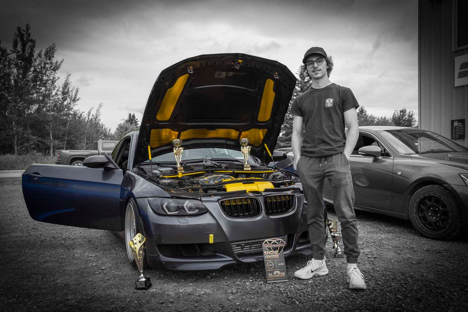 Homme et ses trophées, 
Man posing beside a customized BMW with open hood featuring yellow accents, holding a trophy at an outdoor car show.