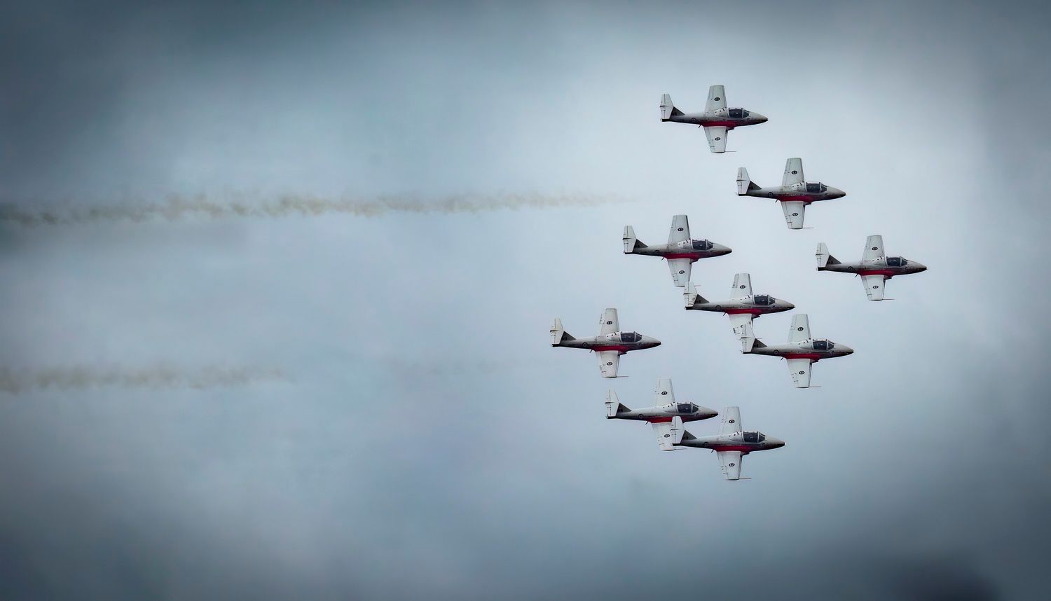 Canadian Snowbirds aerobatic team flying in close diamond formation leaving white smoke trails against grey sky.