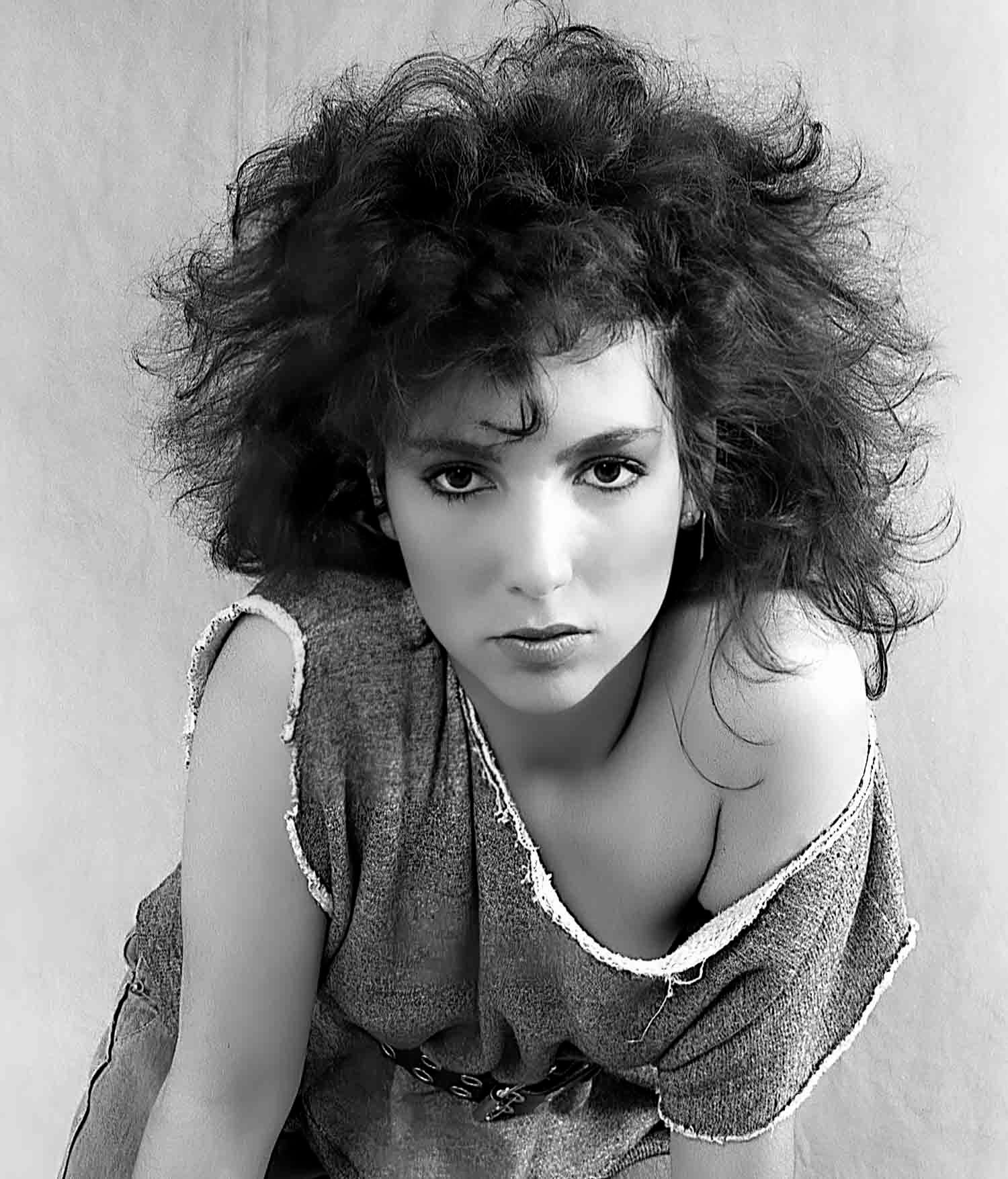 Black and white portrait of a woman with voluminous curly hair wearing a draped top, gazing intensely at camera.