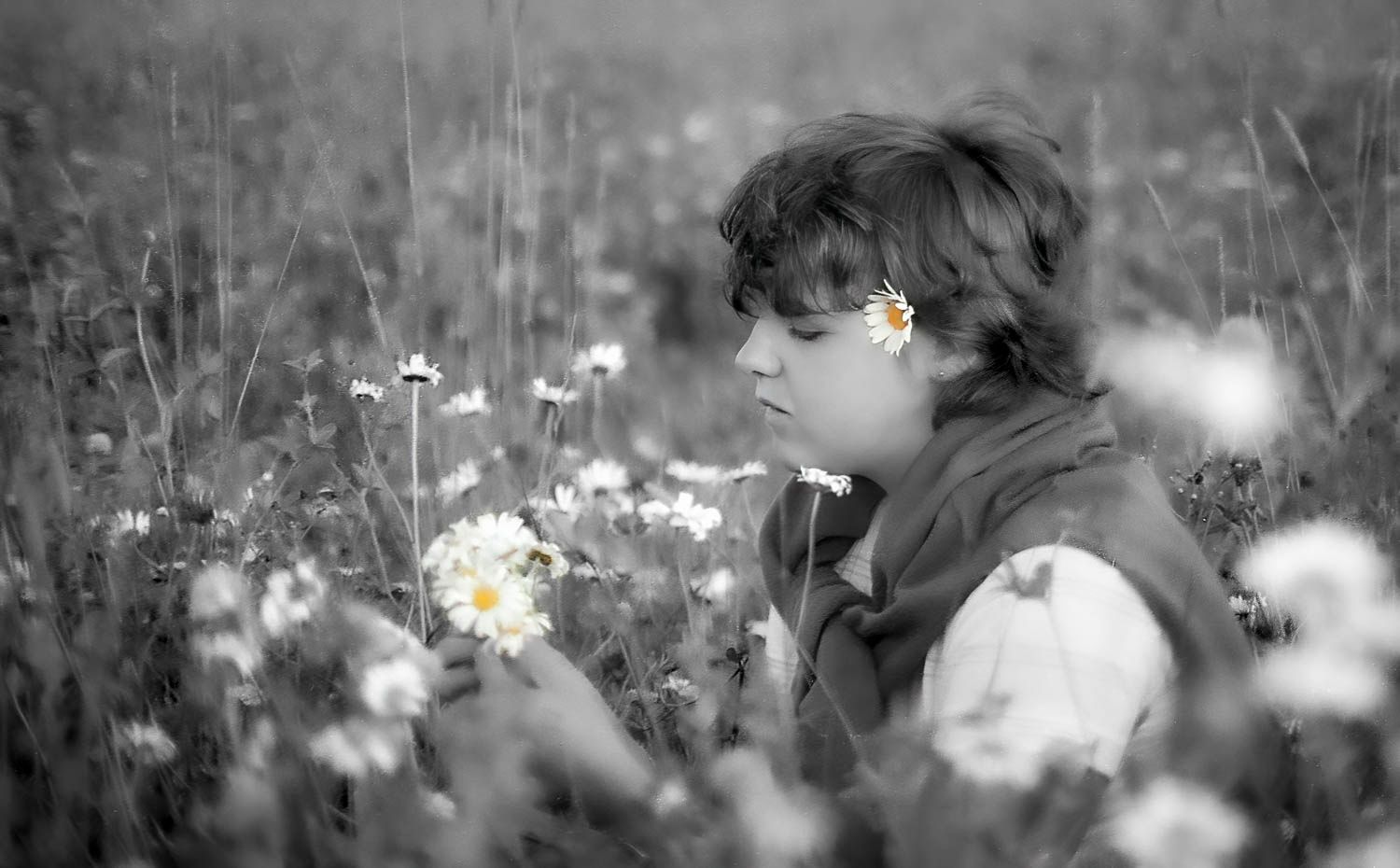 Black and white photo of a child sitting among daisy flowers in a field, looking down thoughtfully.