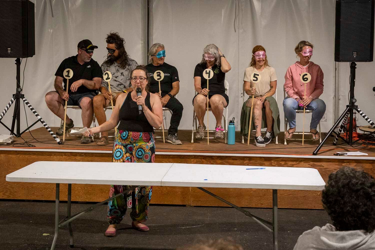 People holding numbered paddles sit on stage during a judging event with a host standing at a table in the foreground.