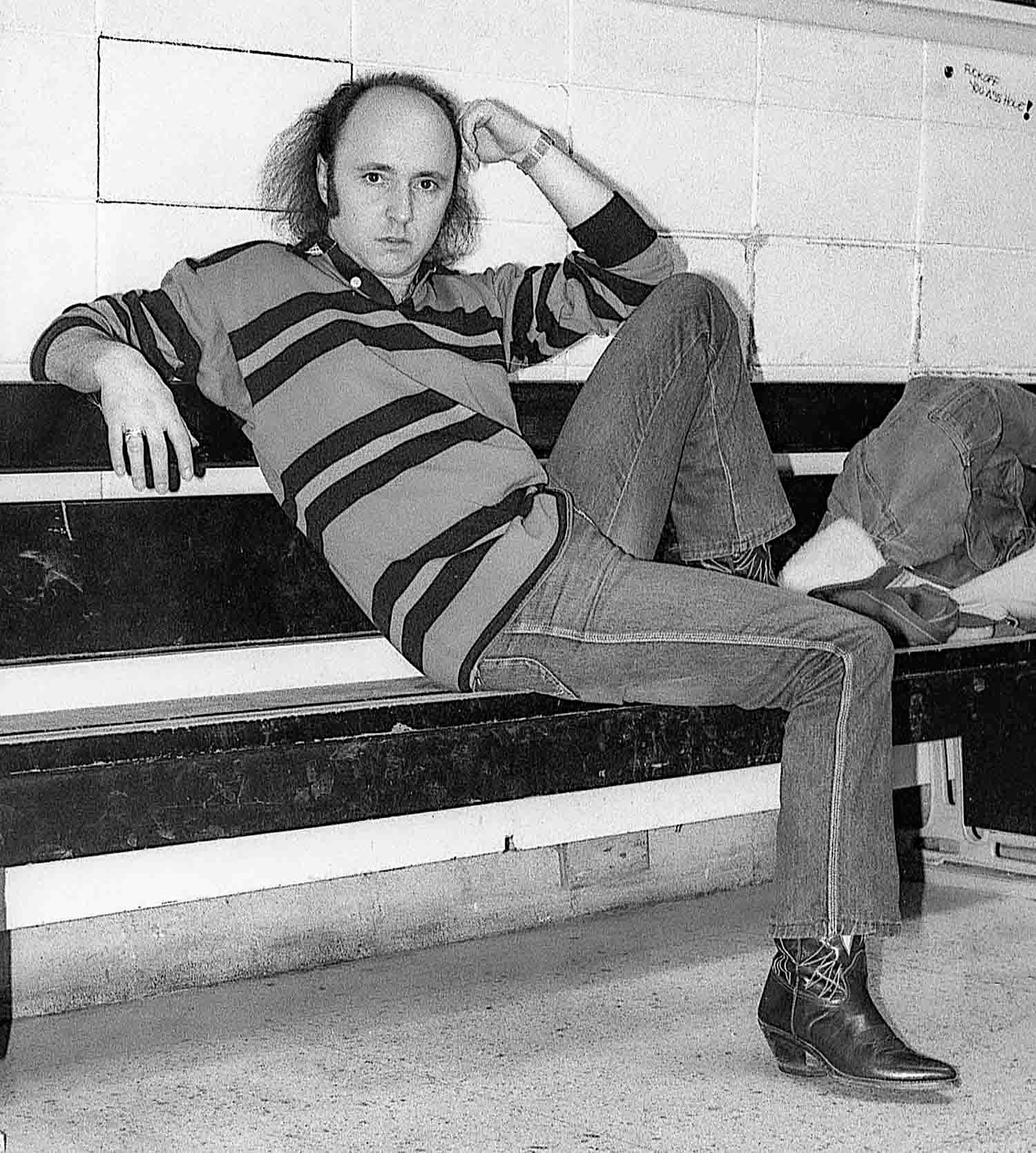 Lucien Francoeur assis sur un banc.
Black and white photo of a man in striped sweater and jeans reclining on bleachers with cowboy boots.