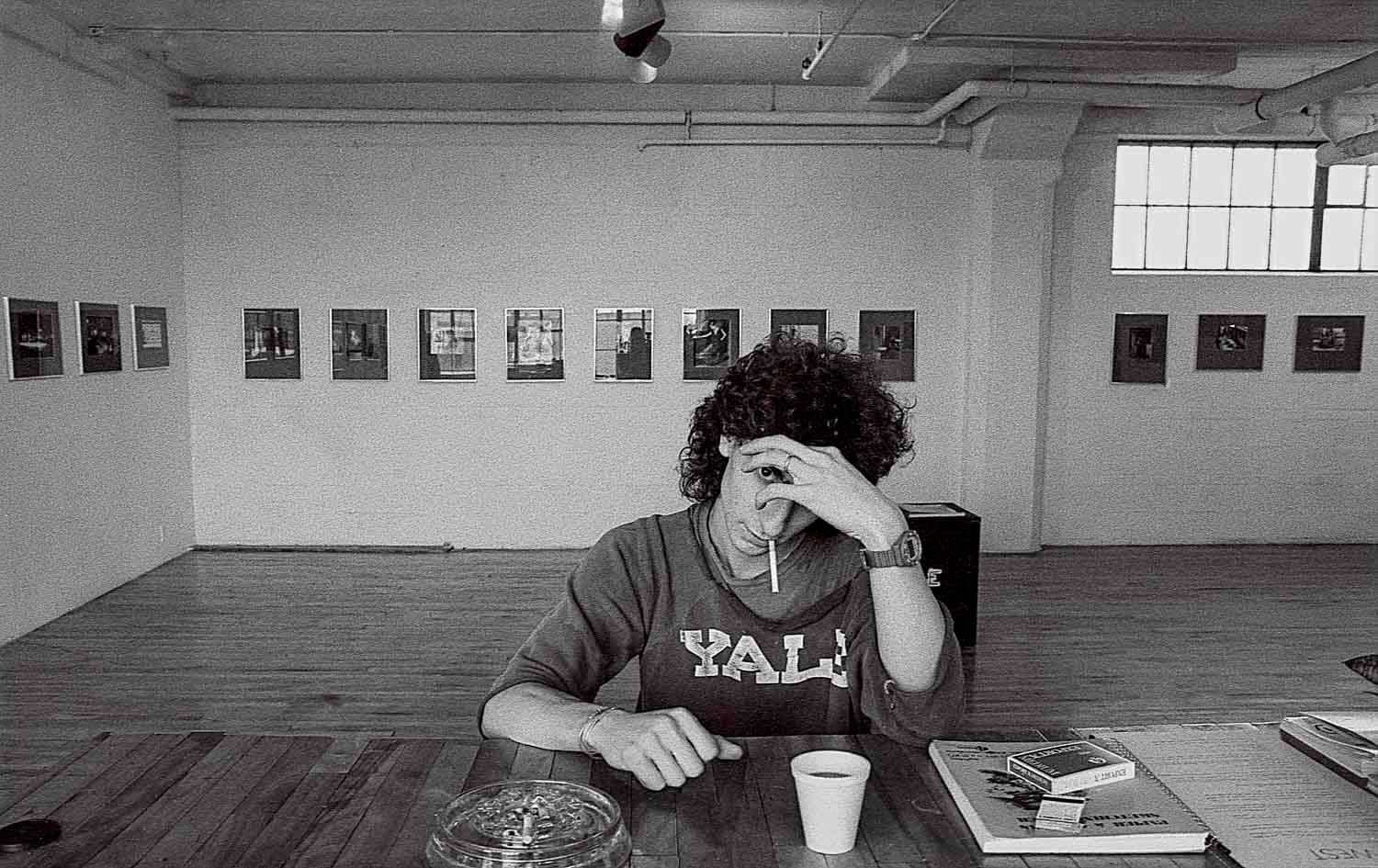 Black and white photo of a person sitting at a wooden table in an art gallery with framed photos on the wall.