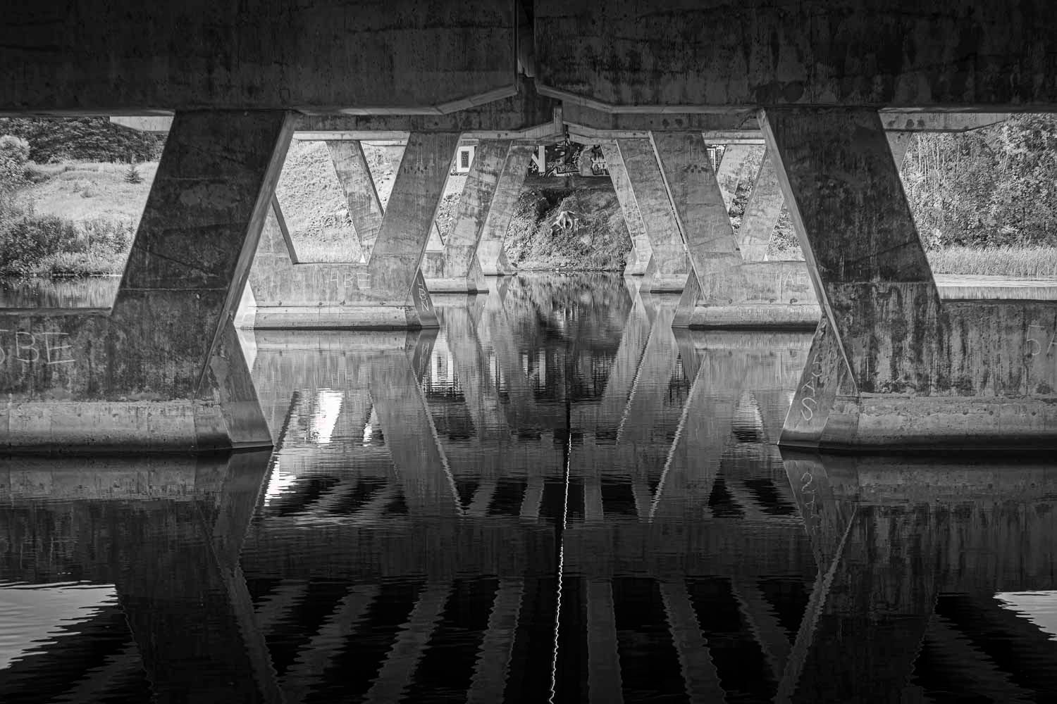 Black and white photo of a concrete bridge underside reflecting symmetrically in still water below.
