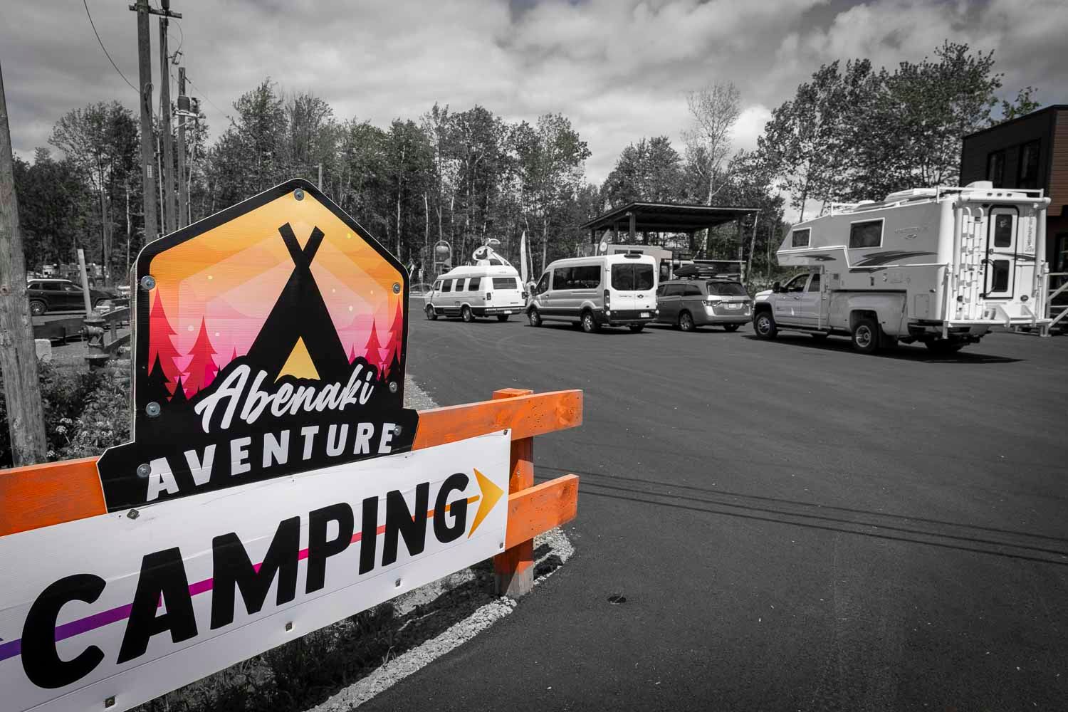 Abenaki Aventure Camping sign in orange with RVs parked in background, black and white nature setting.