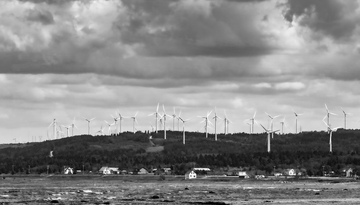 Éole et ses disciples
Cap-Chat, 
Black and white photo of wind turbines on a hilltop above a small coastal town under dramatic cloudy skies.