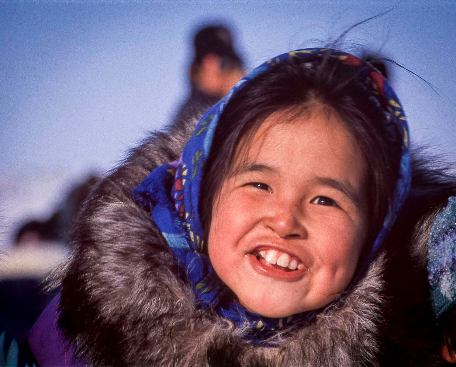 Smiling young Inuit girl wearing a colorful headscarf and fur-trimmed parka in a cold arctic environment.