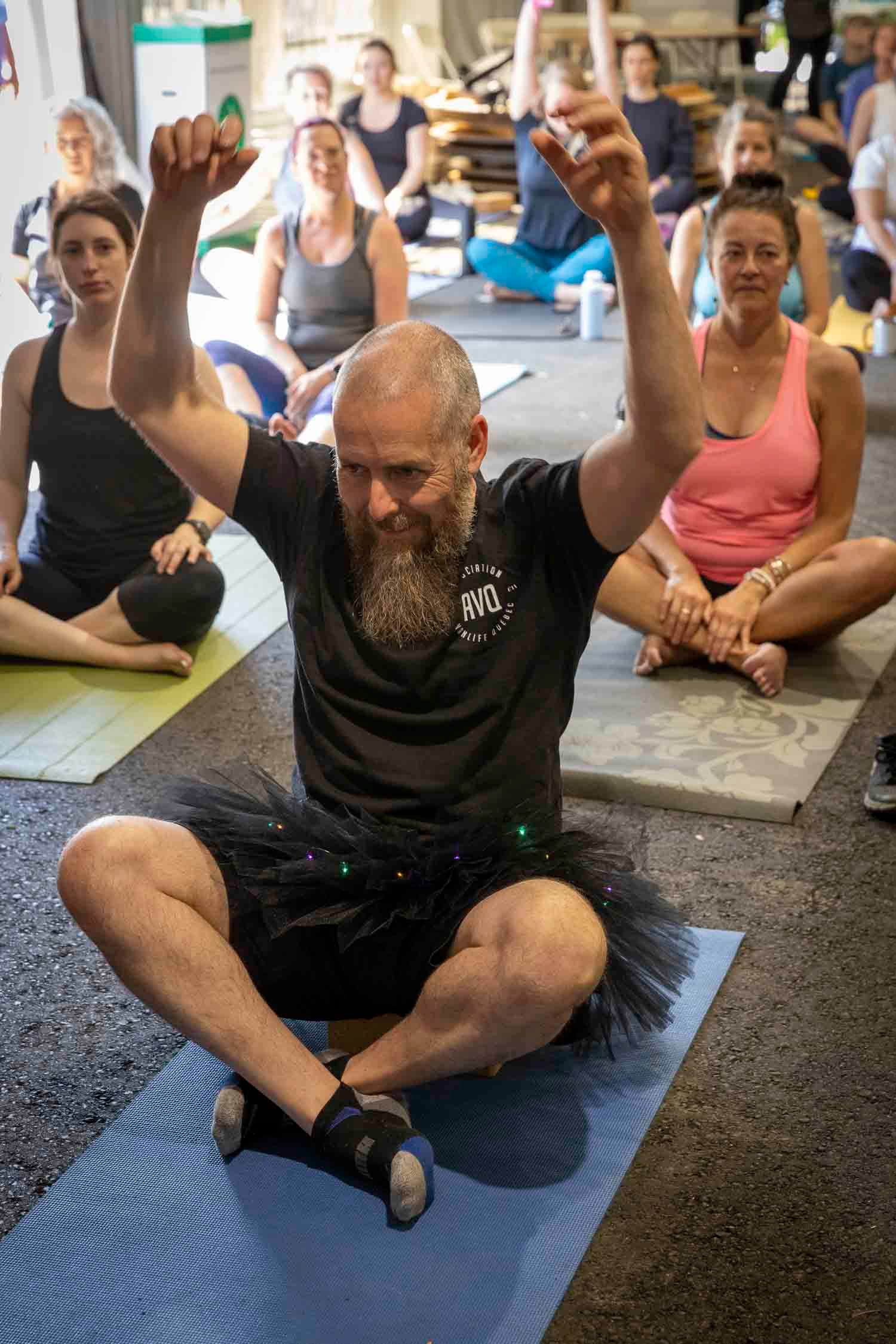 Bald bearded man in tutu sits cross-legged on yoga mat with arms raised leading group fitness class.