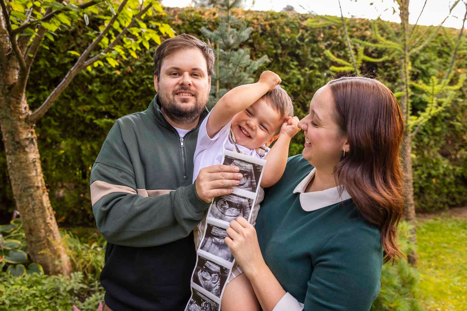 Happy family with young child holding ultrasound photos outdoors, announcing a new pregnancy in a garden setting.