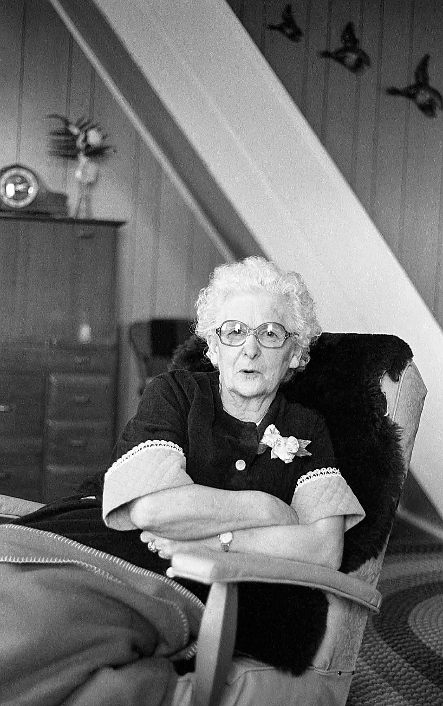 Lauretta.
Elderly woman with white curly hair and glasses sitting in an armchair, arms crossed, in a vintage black and white photo.