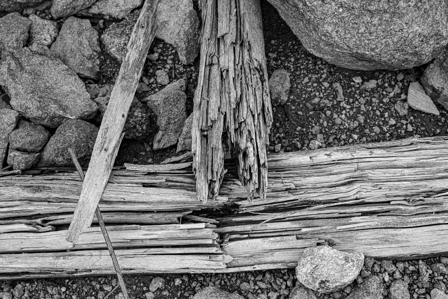 Close-up black and white photo of weathered, splintered driftwood planks scattered among rocks on a shoreline.
