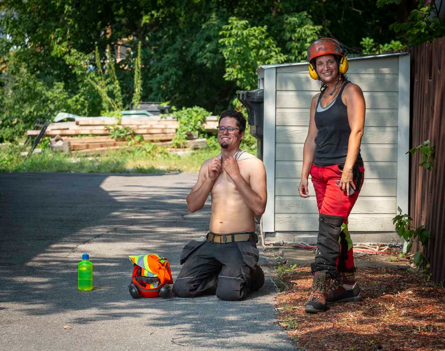 Two workers in safety gear kneel and stand outdoors, one shirtless, near tools and a colorful toy on a sunny driveway.