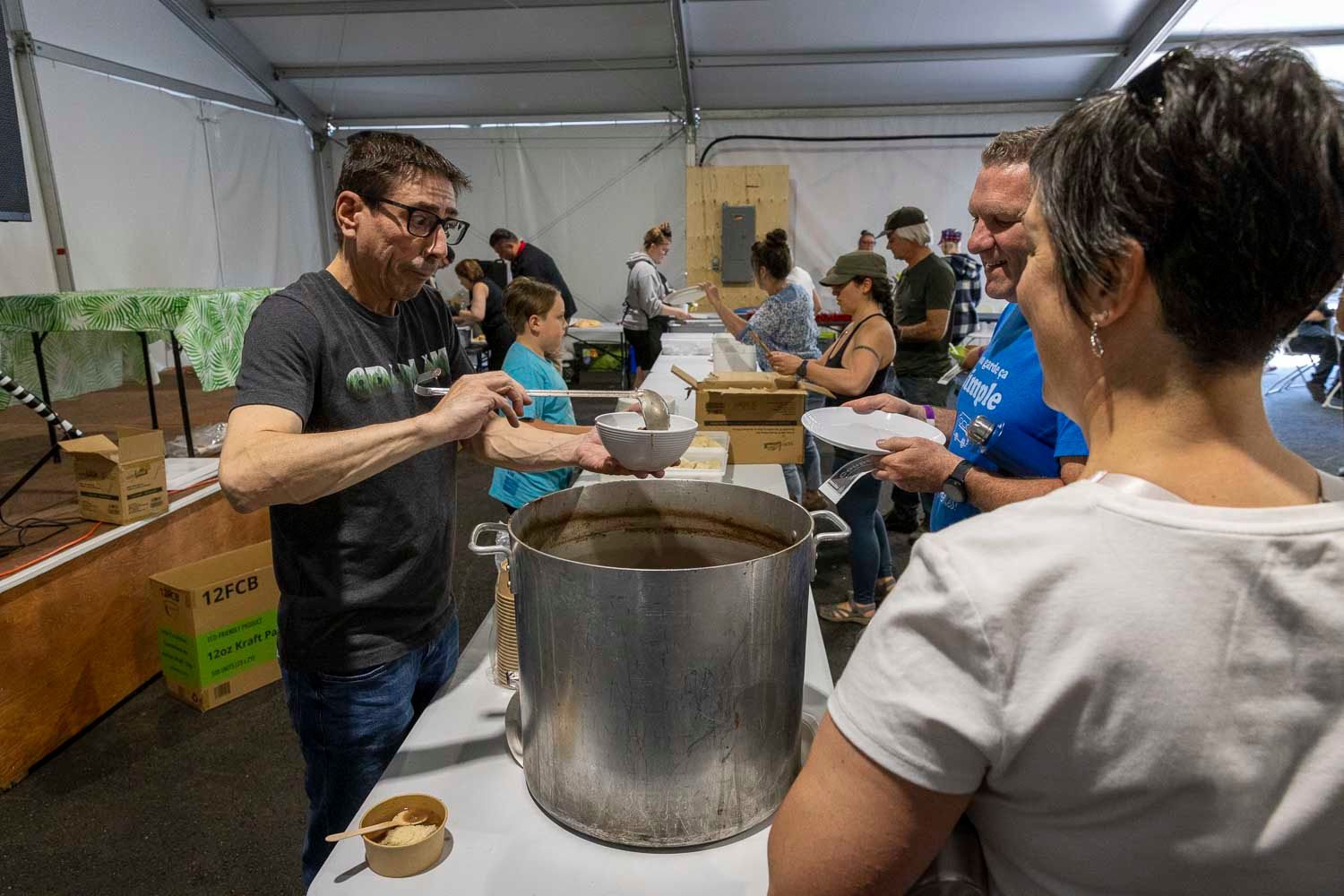 Man serves food from a large stockpot to guests at an indoor food event or community gathering.