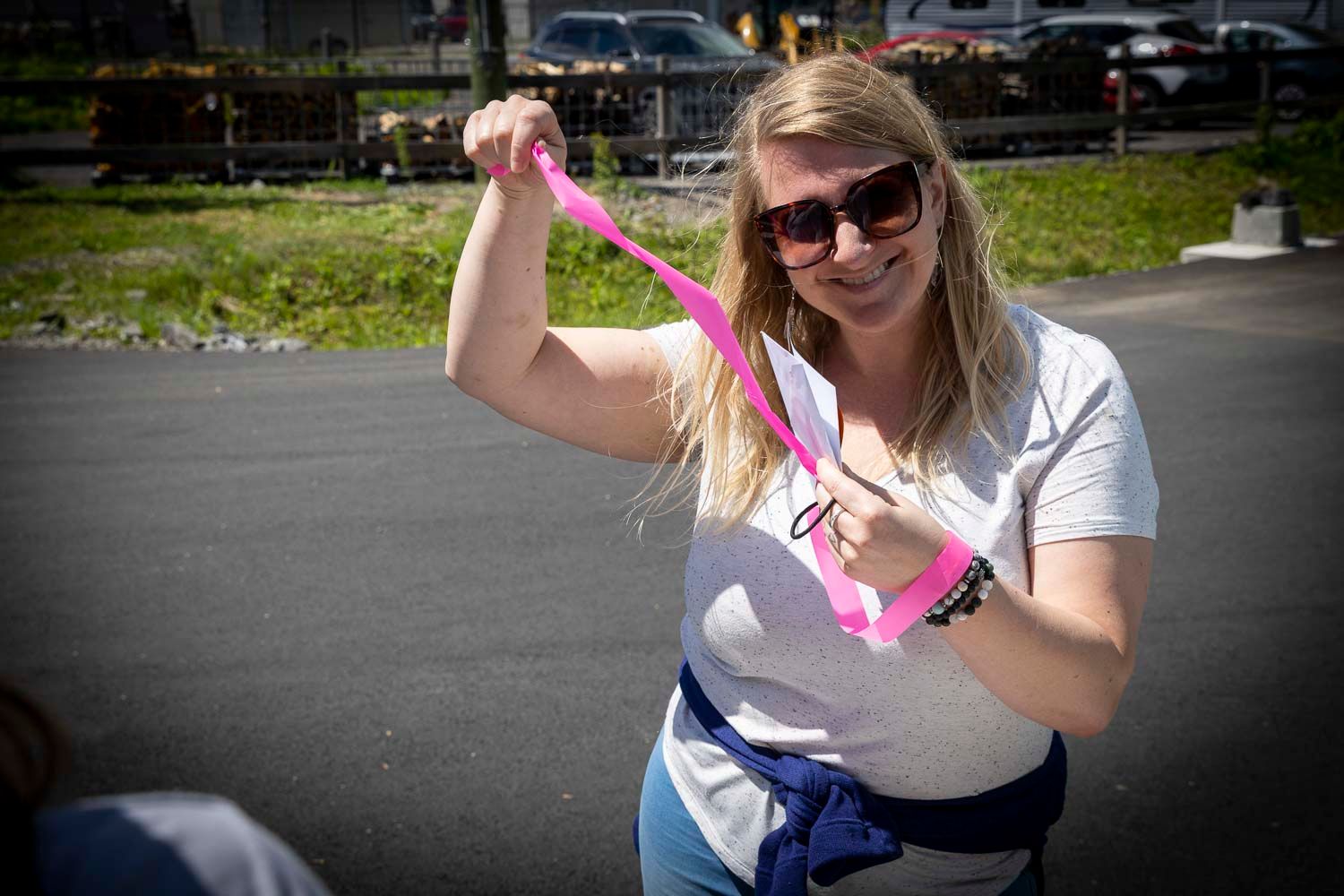 Smiling blonde woman in sunglasses holding a pink ribbon outdoors on a sunny day.