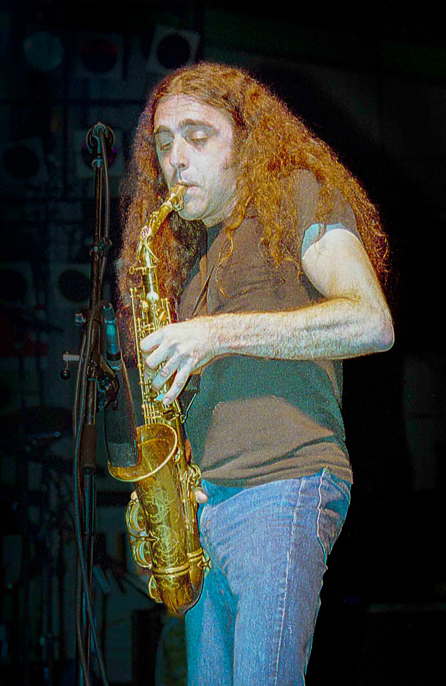Curly red-haired musician playing alto saxophone on stage under colorful concert lights.