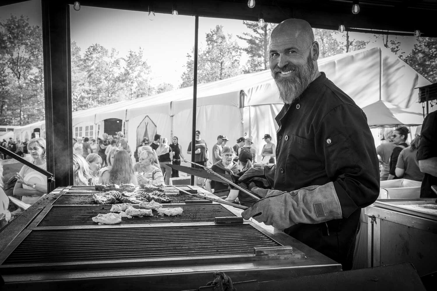 Smiling bald man with beard grilling meat at outdoor BBQ food festival event in black and white photo.
