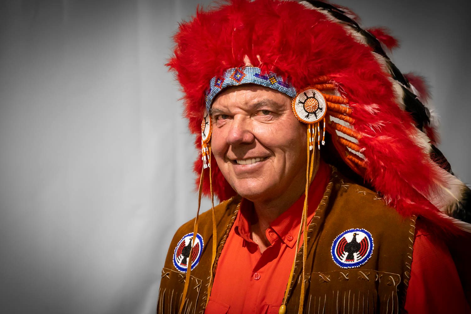 Man smiling in traditional Native American headdress with red feathers and beaded regalia.