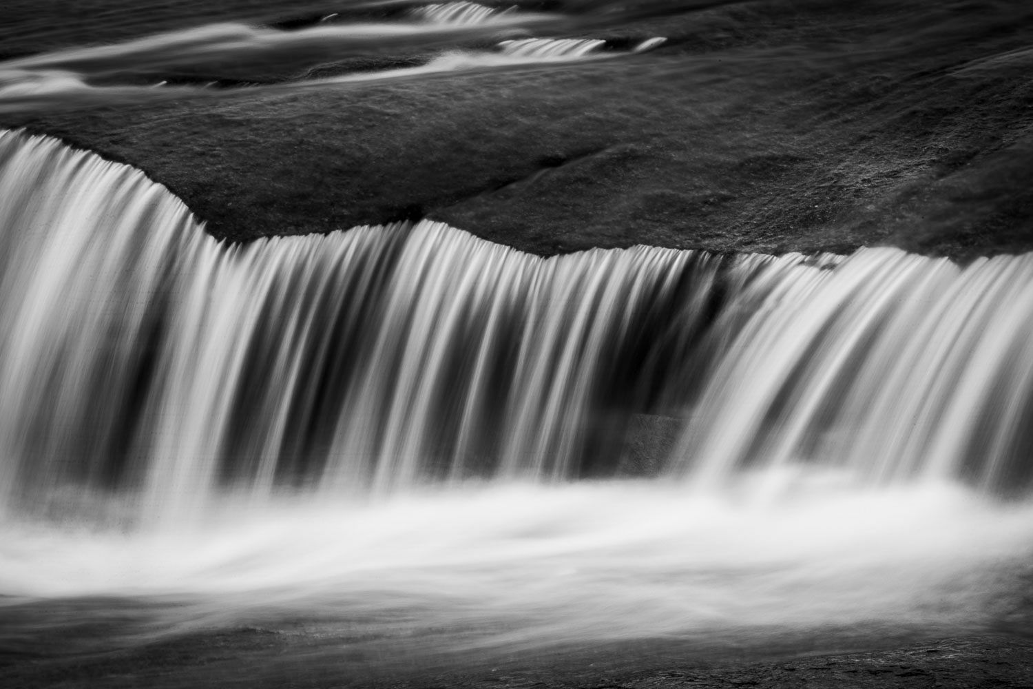 Black and white long exposure waterfall cascading smoothly over dark rocky ledge with silky water motion blur.