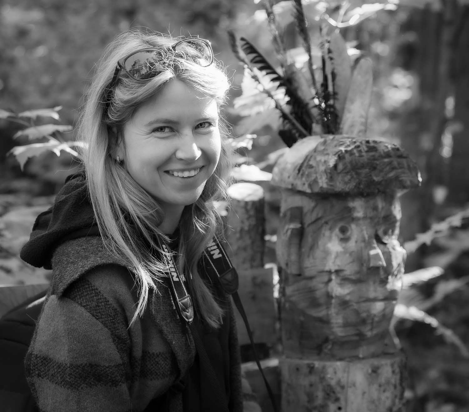 Smiling young woman with blonde hair posing outdoors near a mossy stone pillar in a forest setting.
