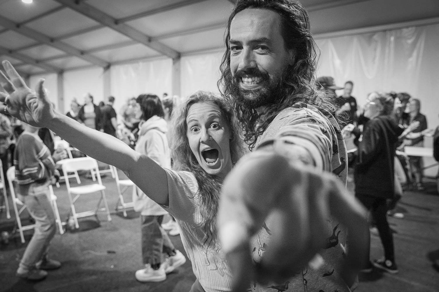 Two excited festival-goers pose energetically at an indoor event, pointing at camera in black and white photo.