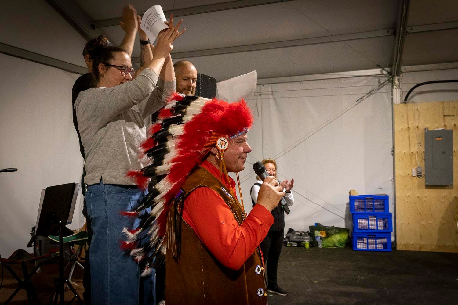 Indigenous chief in traditional feathered headdress speaking at a microphone inside a tent event.