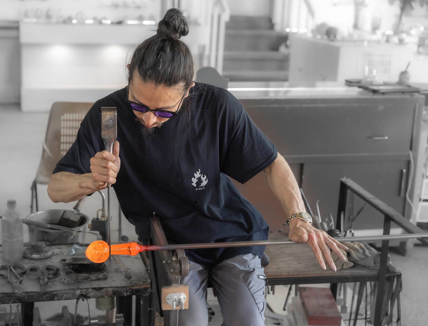 Skilled glassblower shaping molten orange glass with tools at a workbench in a professional studio.